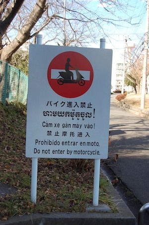 バイク進入禁止の看板。日本語のほか、ラオス語、ベトナム語、中国語、スペイン語、英語の6か国語で書かれている。ゴミ捨ての看板なども、多国籍な言語で表示