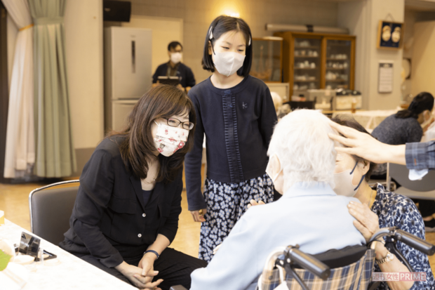 〇〇ちゃんと高齢者の名前を呼んで笑顔を引き出し、緊張を一瞬でほぐすかづきさん。万博やボランティアには孫(右隣)が見学に訪れ、その仕事ぶりを「カッコいい」と絶賛(撮影/矢島泰輔)